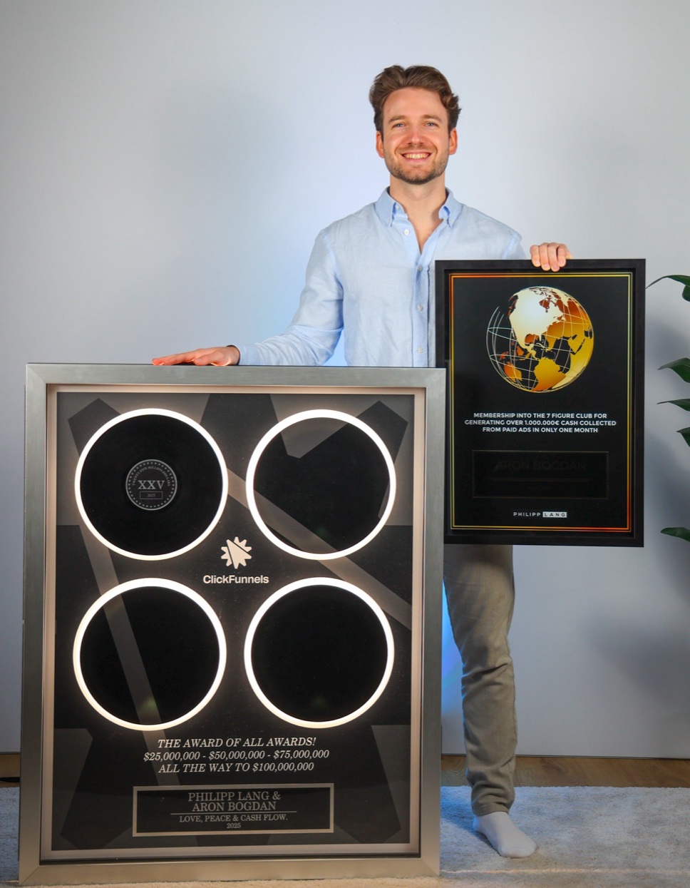 Aron Bogdan holding both of his awards: the large ClickFunnels 2-Comma-Club-C award for generating over $25M from a single funnel, and the 7 Figure Club award for over €1M cash collected from paid ads in a single month.